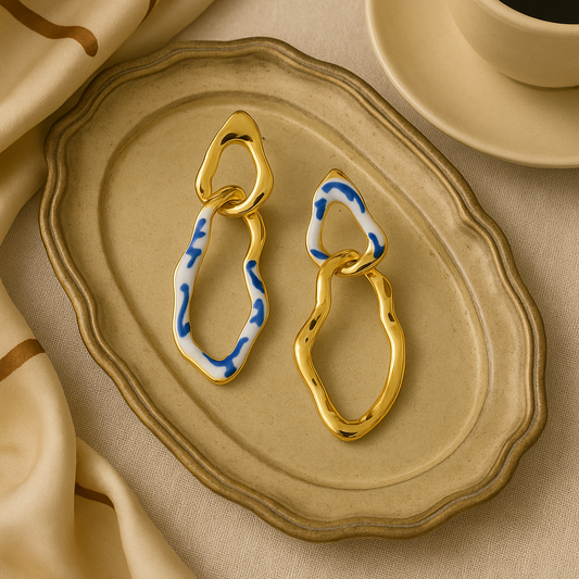 A pair of gold-colored dangle earrings with blue and white floral patterns, displayed on a round beige plate.