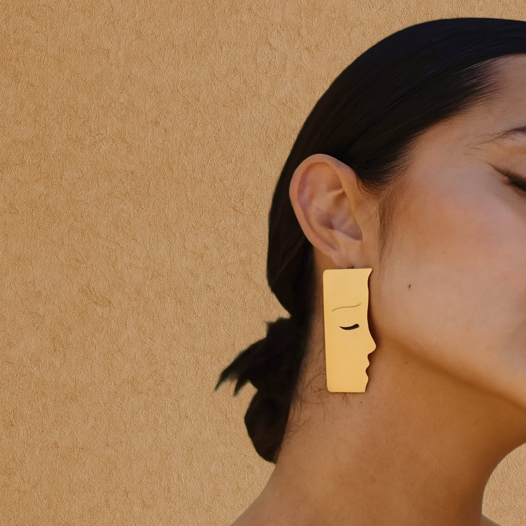 Gold earrings with a face design worn by a person against a beige background